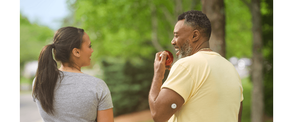 A man walking outdoors and eating an apple, he has a visible sensor on his arm 