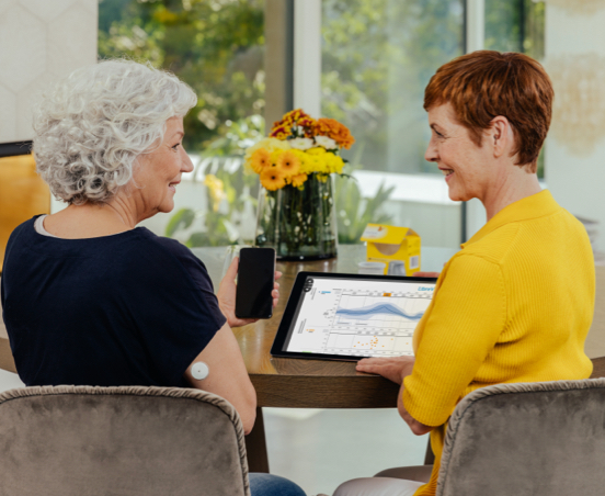 A Healthcare Professional and a patient are discussing the patient's glucose data on a tablet screen. The patient has a sensor visible on the back of her upper arm and is holder her phone.