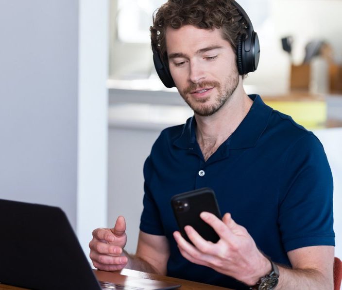 Man with headphones sitting in front of a laptop looking at his phone